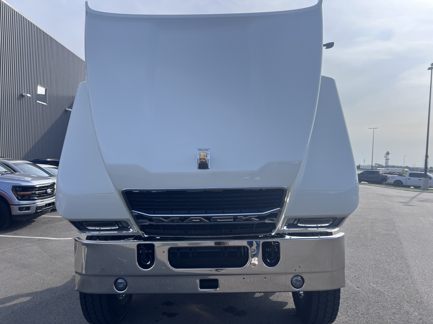 Front view of a 2026 Mack PI64T truck cab with hood raised revealing grille, bumper, and dual fog lights.