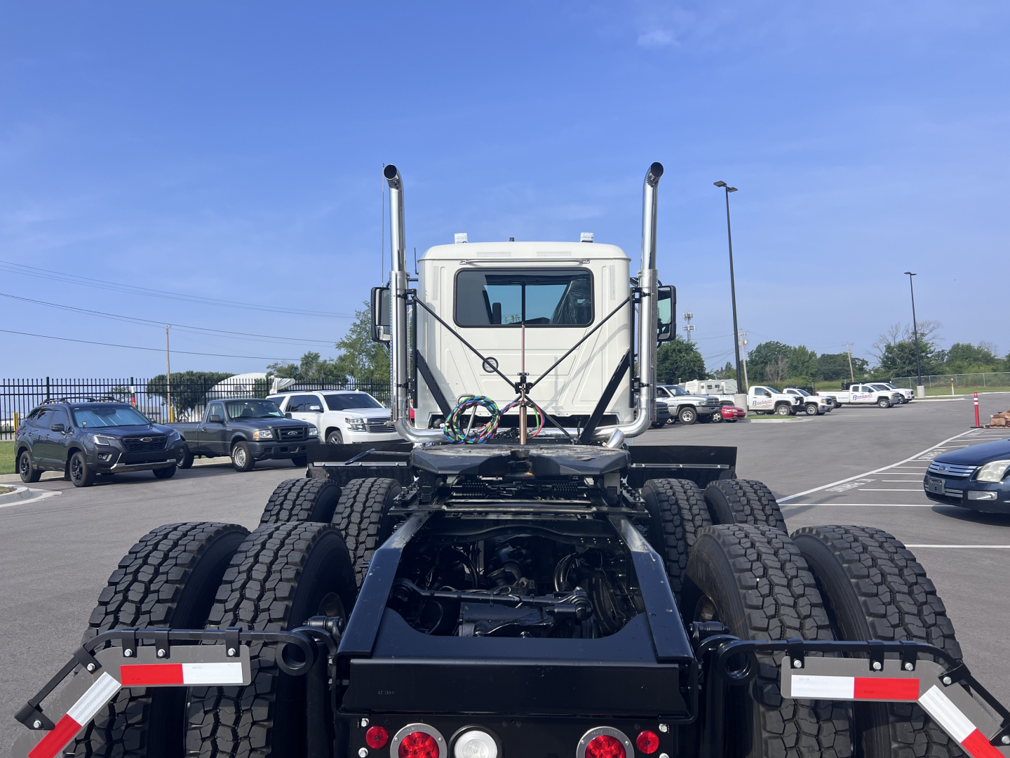 Truck chassis rear view of a new Mack PI64T with dual rear axles, fifth wheel, and dual stacks, showing heavy-duty tires and undercarriage components.