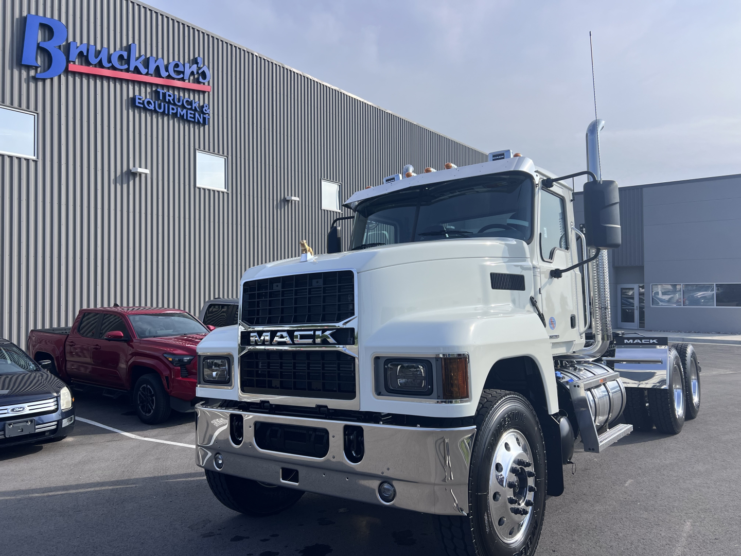 Mack PI64T truck in white, front-quarter view, showing grille with MACK, large bumper, side mirrors, dual rear wheels, chrome steps; Bruckners Truck Equipment building in background.