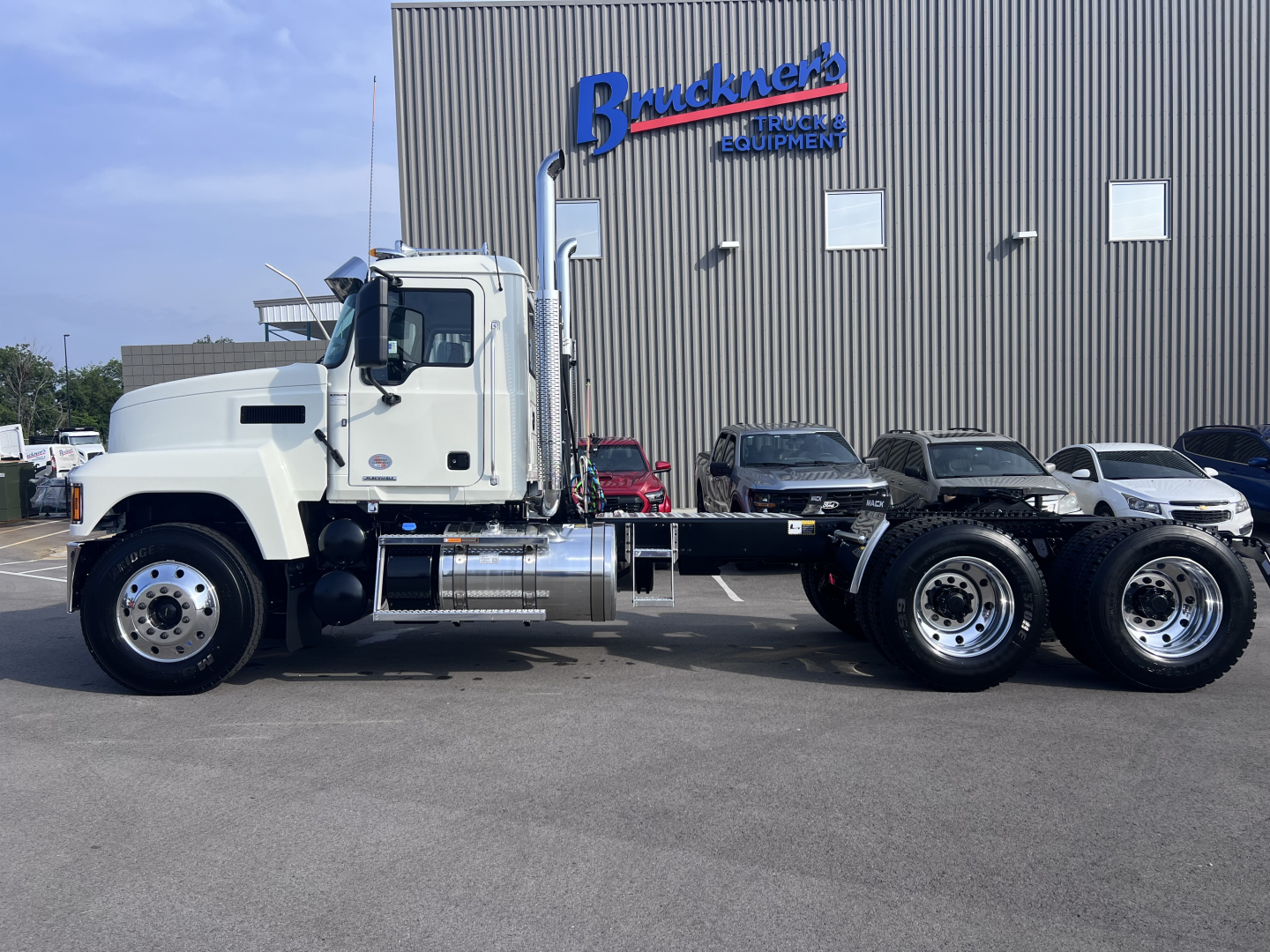 Mack PI64T conventional daycab truck with white cab and dual rear axles, parked in front of a building with 'Bruchne...' sign; side profile shows chrome fuel tanks and exhaust stack.