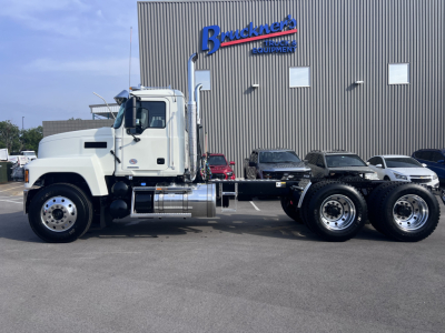 Mack PI64T conventional daycab truck with white cab and dual rear axles, parked in front of a building with 'Bruchne...' sign; side profile shows chrome fuel tanks and exhaust stack.