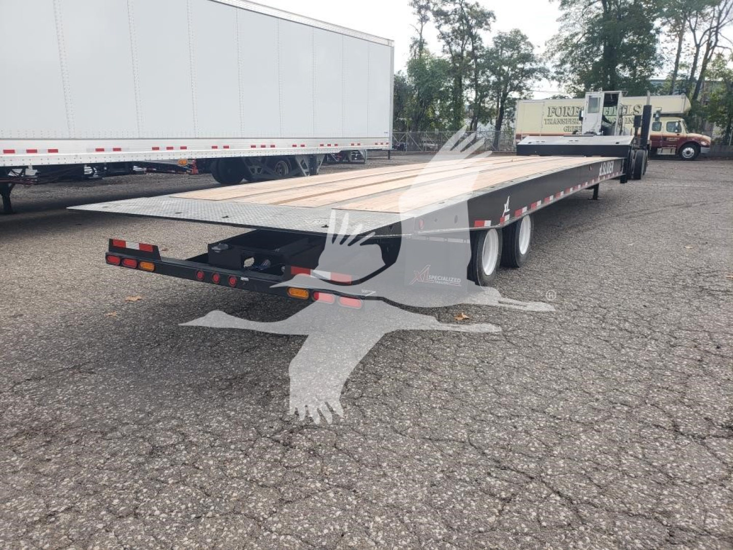 Flatbed trailer with rear hitch and dual rear axles. Visible text includes none clearly readable from distance; truck yard in background.