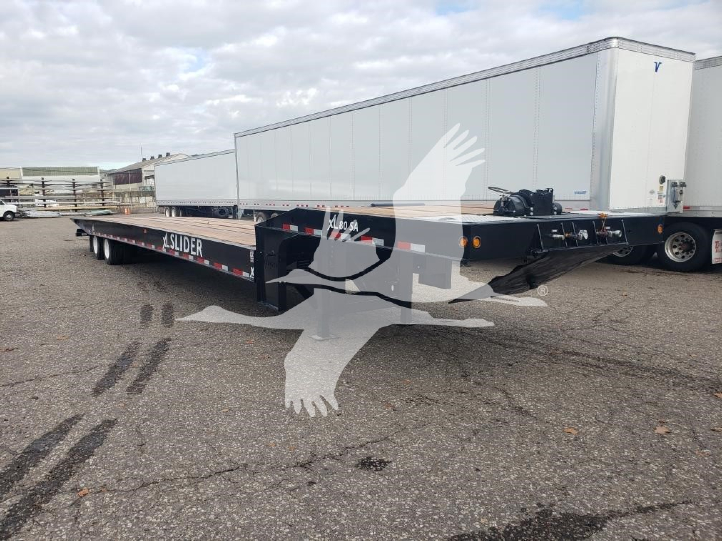 XL Specialized XL80SA traveling axle trailer with flatbed and low deck in a parking lot; side shows 'XL SLIDER' branding and a white trailer in background.