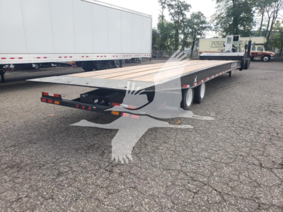 Flatbed trailer with rear hitch and dual rear axles. Visible text includes none clearly readable from distance; truck yard in background.
