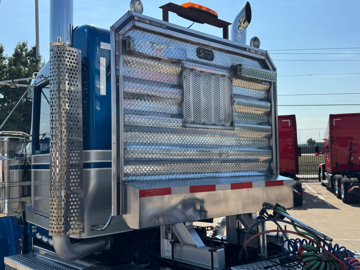 Kenworth W900L rear/side view showing diamond plate toolbox and exhaust stack on blue conventional daycab truck.
