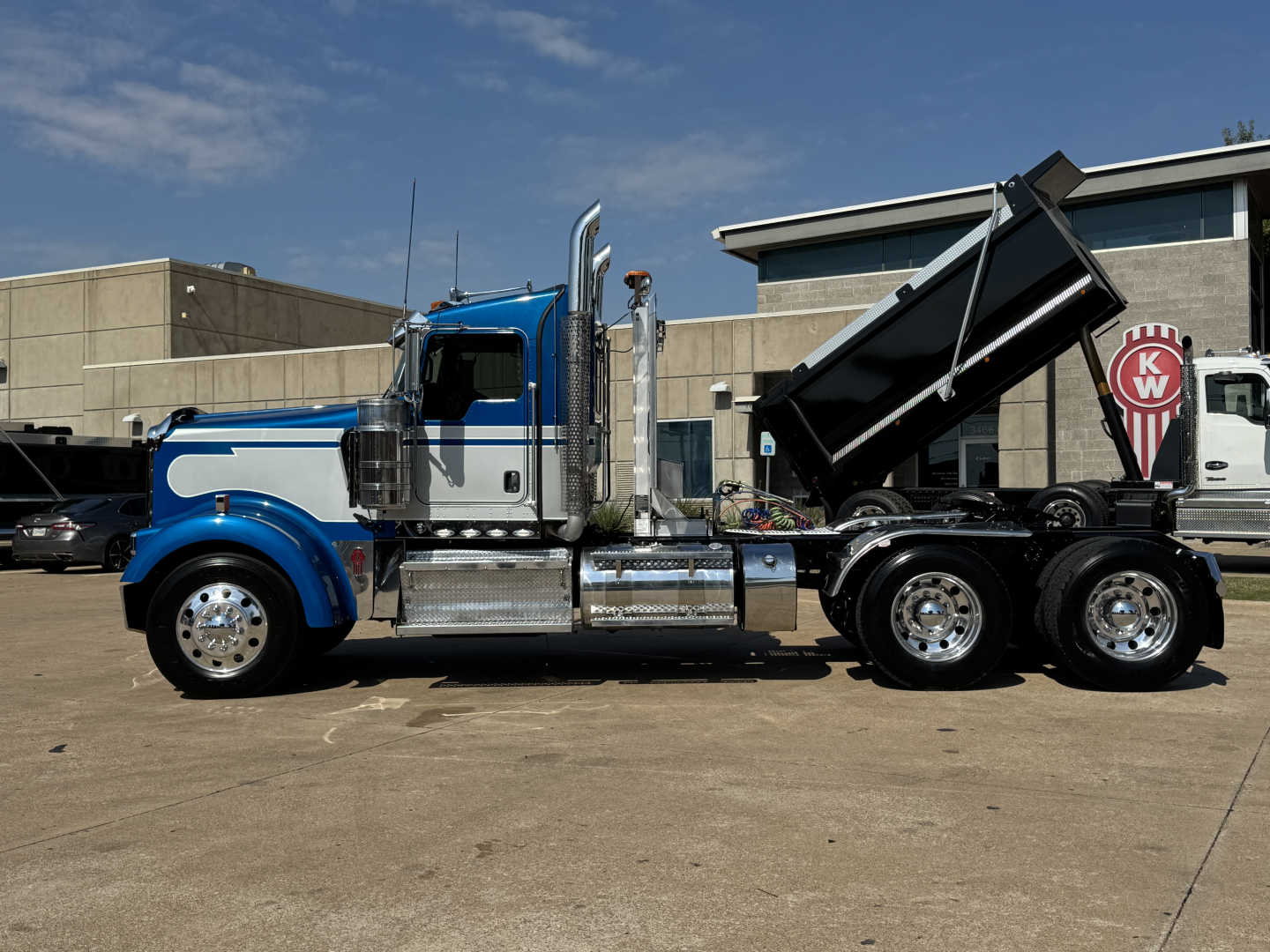 Blue Kenworth W900L conventional daycab truck side view showing dump bed in raised position and chrome exhaust stacks.