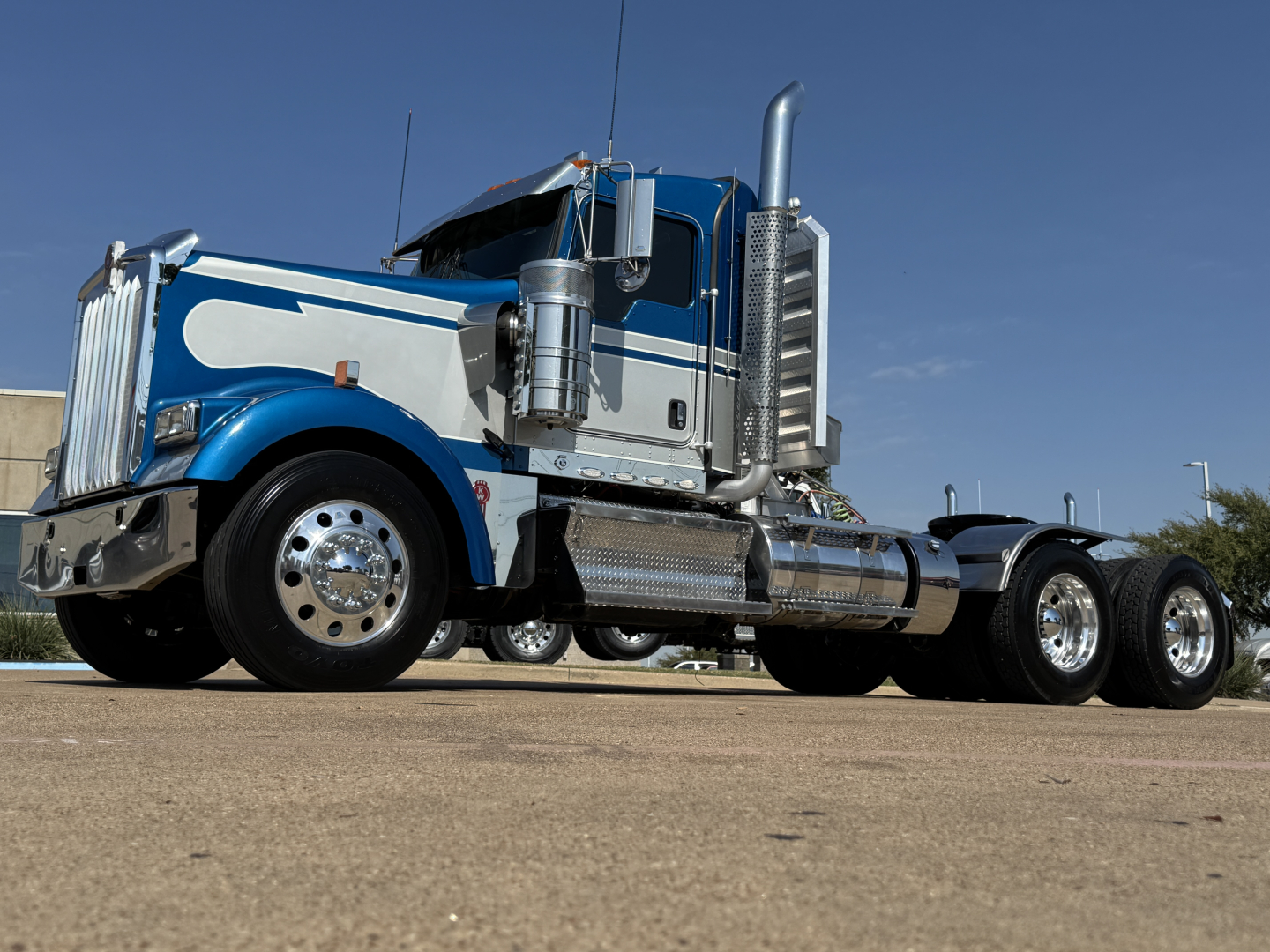 2024 Kenworth W900L truck in blue and white with chrome exhaust stacks, front chrome bumper, and dual rear axles.