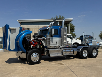 Kenworth W900L conventional daycab truck on a lot with chrome fuel tanks, blue air intake, and dual rear axles; engine bay exposed on the left side.