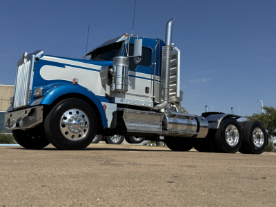 2024 Kenworth W900L truck in blue and white with chrome exhaust stacks, front chrome bumper, and dual rear axles.