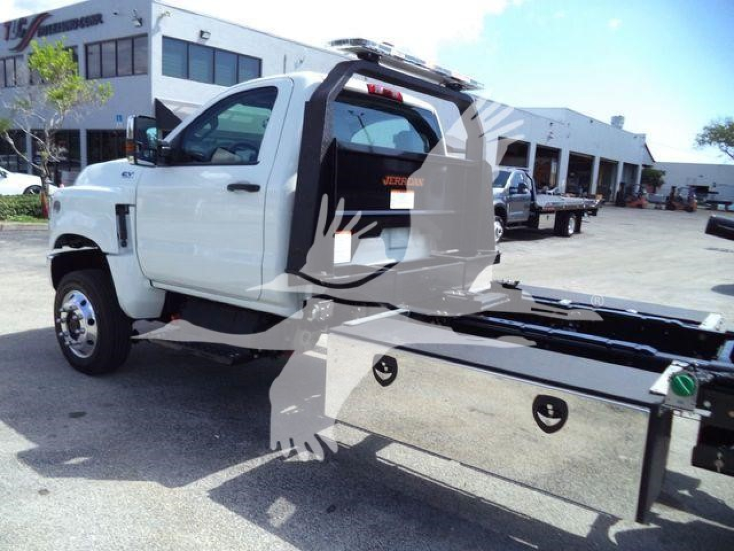 Truck cab with rollback carrier system mounted, white exterior, side view showing cab and towing platform, dealership lot in background.
