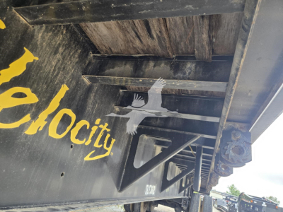 Flatbed trailer side with yellow logo and metal frame, underside view showing structural crossbeams and hitch area.