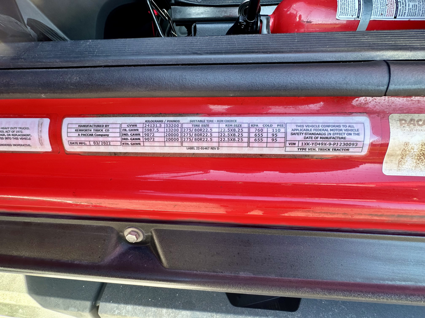 Vehicle data sticker on a red Kenworth T680 door jamb with VIN 1XKYD49X9PJ230093 and stock number 0632224, showing standard load specs and compliance markings.