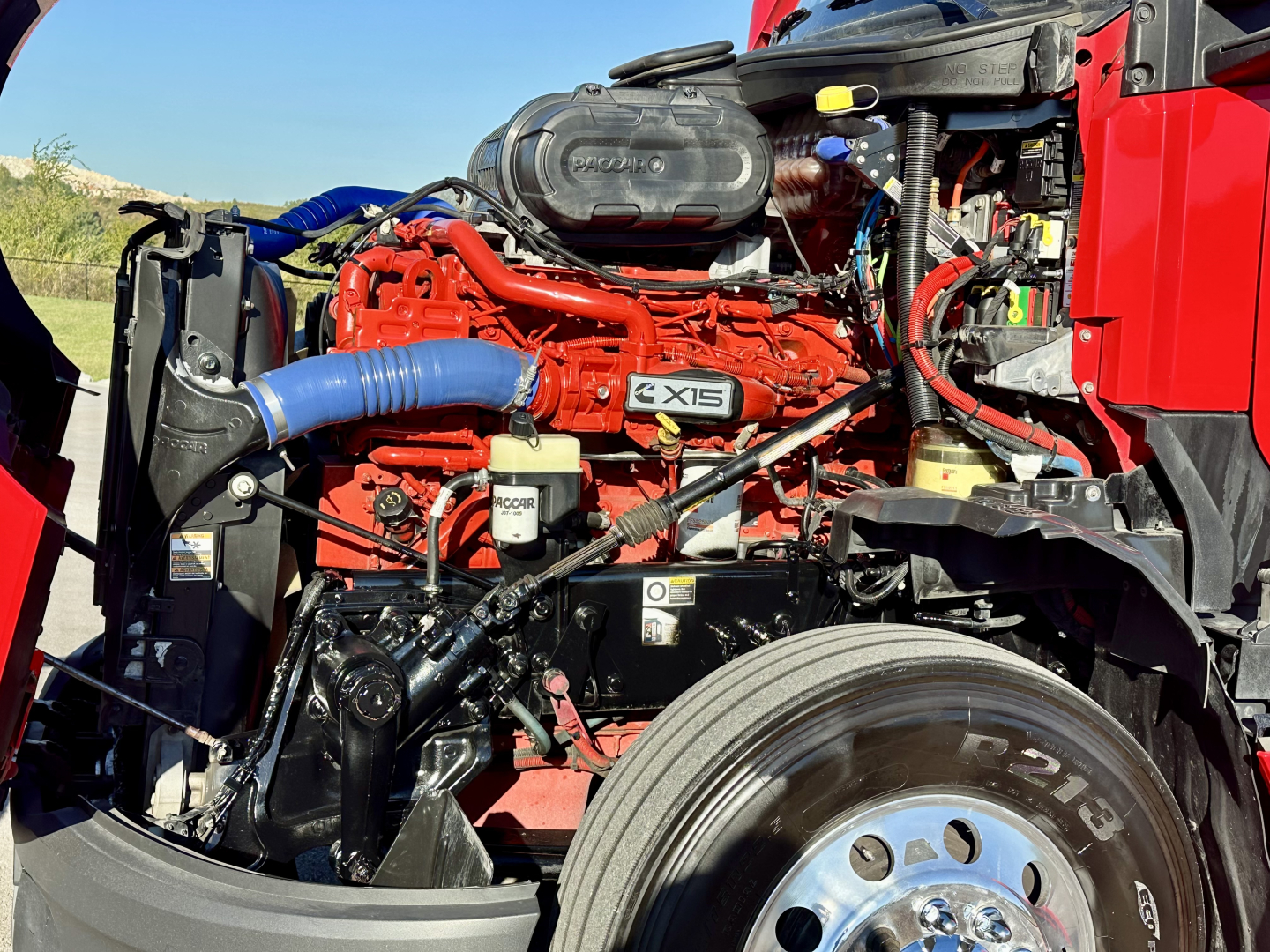 Kenworth T680 engine bay showing 6X18 engine badge, red diesel engine with blue turbo hose, coolant reservoir, air intake, and surrounding components.