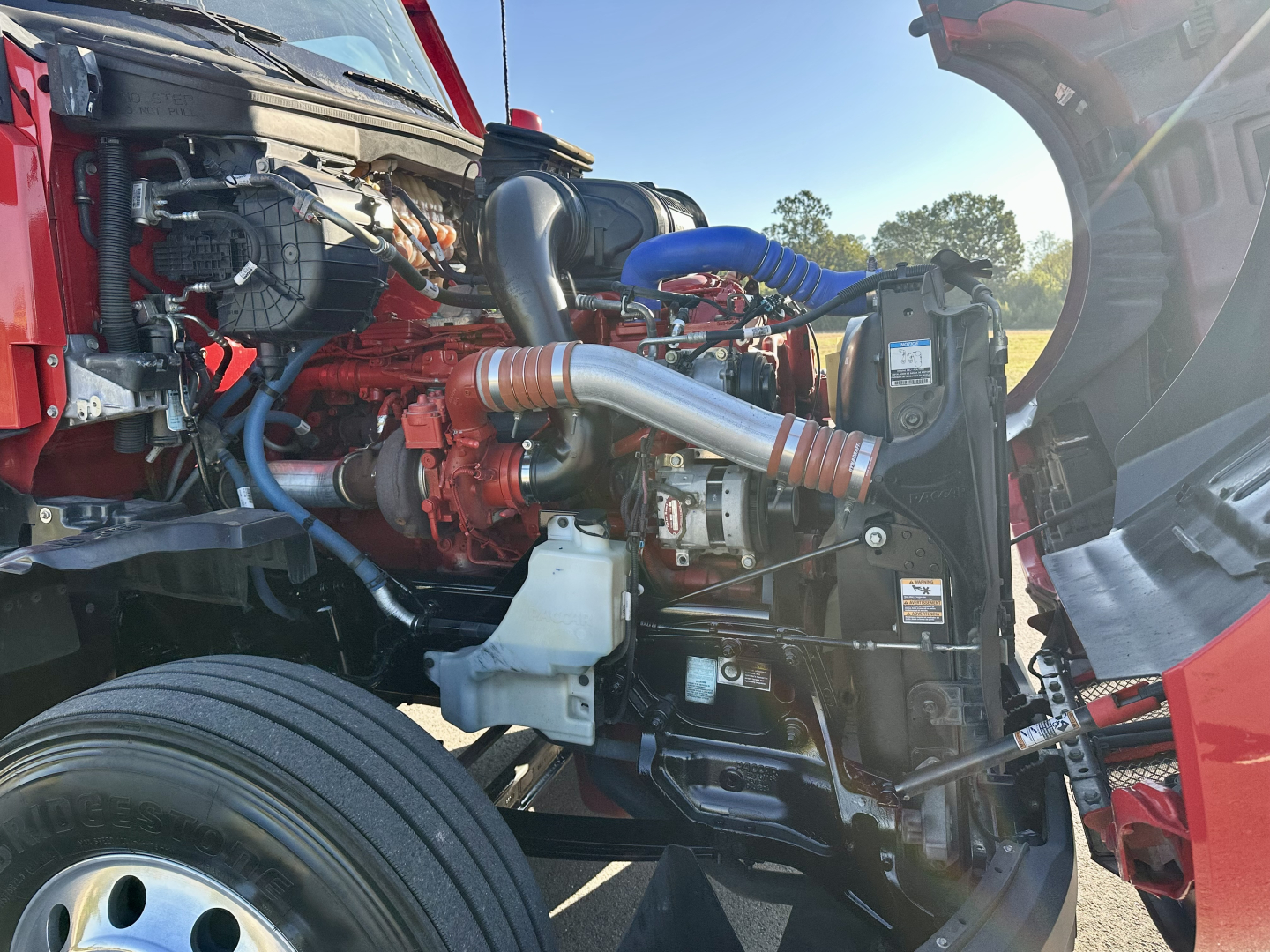 Kenworth T680 engine bay view showing red diesel engine, intercooler piping, blue hose, air intake and radiator. Visible components include turbocharger pipe, coolant reservoir, and surrounding chassis.