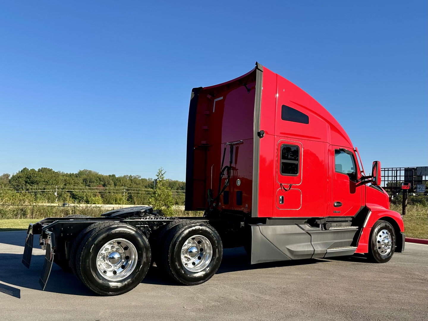 Kenworth T680 semi-truck in red cab, side profile view showing sleeper and dual rear axles, on a paved lot with clear blue sky.