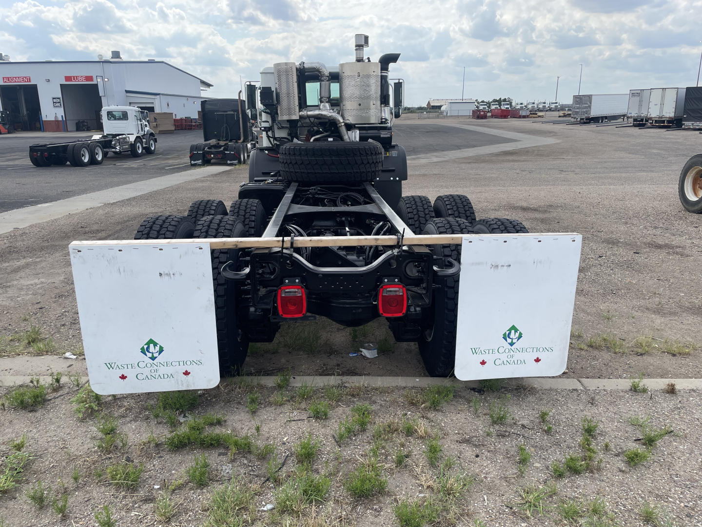 Rear view of a new 2026 Mack TE64 truck chassis showing dual white mud flaps with Waste Connections Canada logos, rear lights, and spare tire mounted between the chassis.