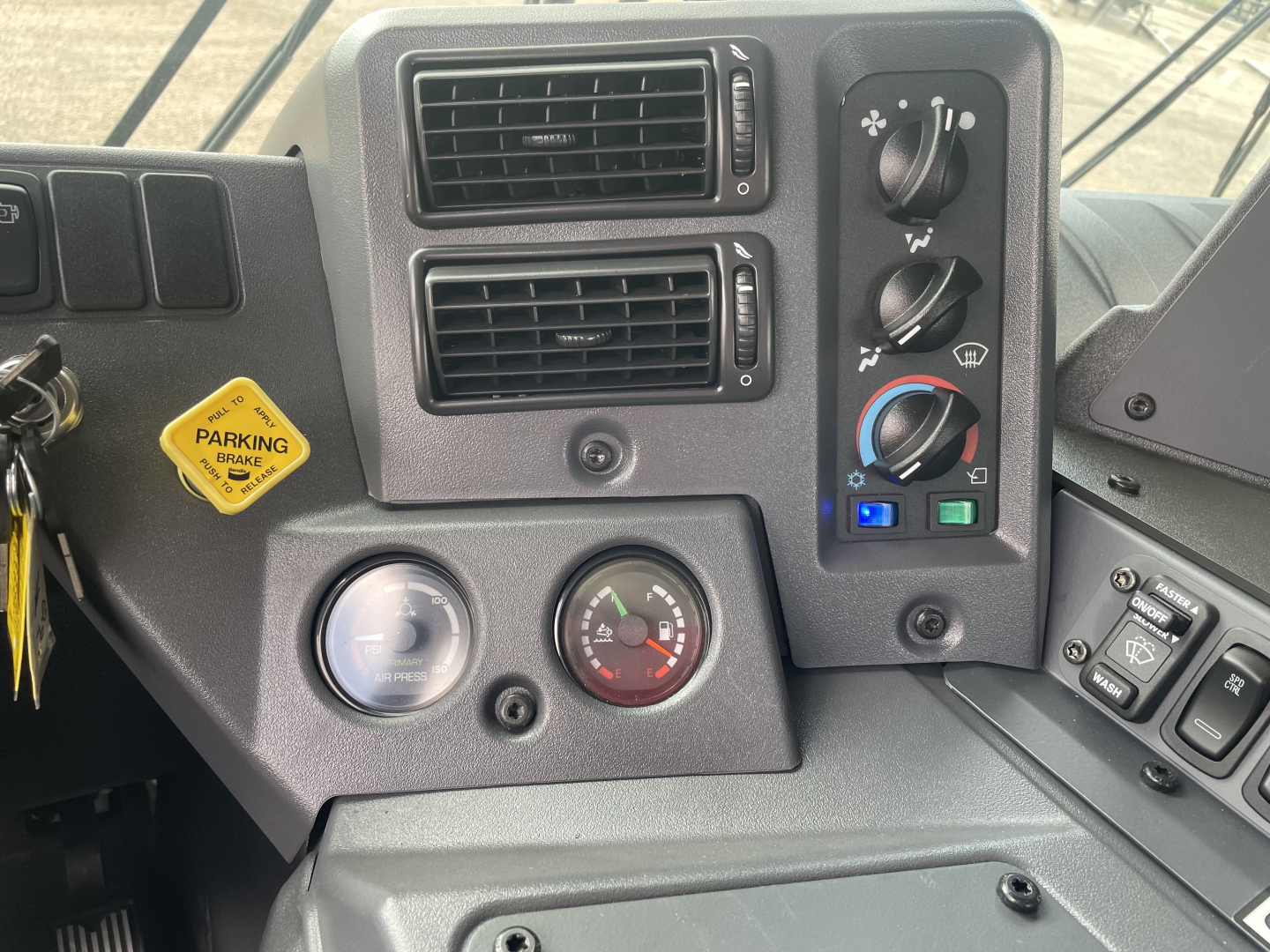 Dashboard of a Mack TE64 truck showing HVAC vents, controls, gauges, and switches with a parking brake sticker and keys in the ignition.