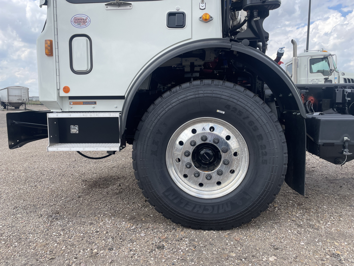 Mack TE64 truck front wheel and fender area showing front tire and wheel well, part of cab with access panel and bumper; visible text on tire sidewall and small badges on cab.