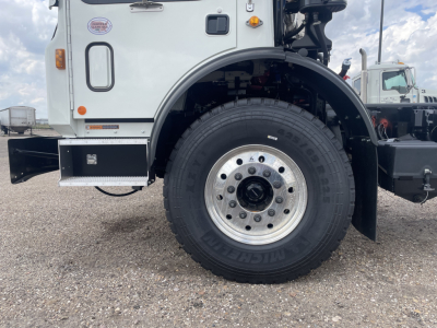 Mack TE64 truck front wheel and fender area showing front tire and wheel well, part of cab with access panel and bumper; visible text on tire sidewall and small badges on cab.