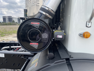 Truck engine bay area featuring a large round air intake canister with warning labels, metal intake pipe, and a small black injector/valve component mounted on the side.