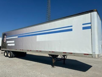 Trailmobile van trailer side view showing long silver body with blue stripes and dual rear wheels on a trailer yard.