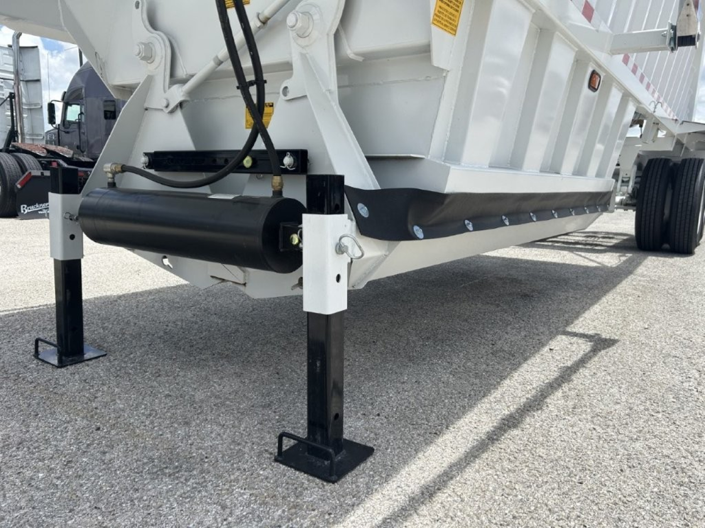Dump trailer undercarriage with white body, black hydraulic cylinder and landing gear leg, showing support A-frame and wheel area.