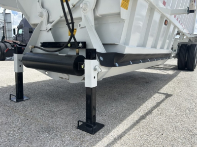 Dump trailer undercarriage with white body, black hydraulic cylinder and landing gear leg, showing support A-frame and wheel area.