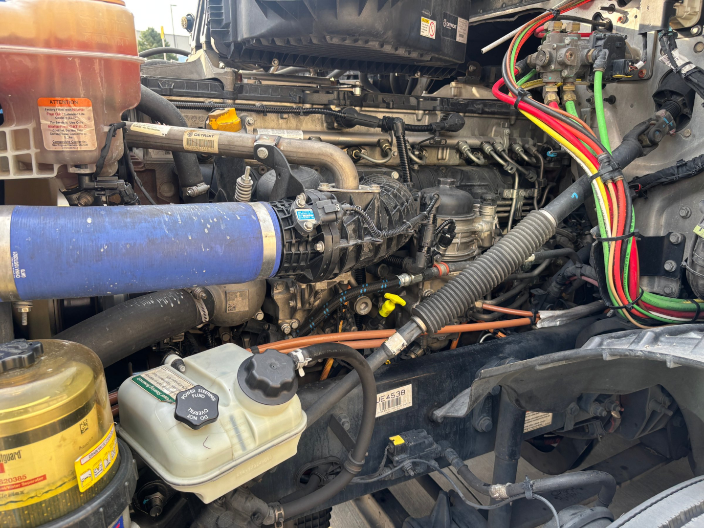 Truck engine bay with various hoses, coolant reservoir, and wiring harness visible in a Freightliner engine compartment.