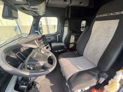 Truck cab interior of a Freightliner Cascadia showing driver's seat and passenger seat, dashboard, steering wheel, center console. The seat headrest reads CASCADIA. Visible text on seat and control area.
