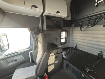 Truck cab interior with sleeper, dash left, storage compartments above, and bunk area; passenger seat visible; overhead cabinets and curtains.