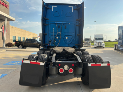 Rear view of a blue Freightliner tractor cab with mud flaps and dual rear axles showing fifth wheel area and back of sleeper with hoses and lights.