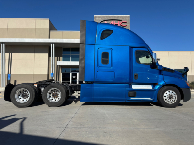 Truck side view showing a blue Freightliner PT126064ST sleeper cab with tandem rear axles, parked in lot at dealership.