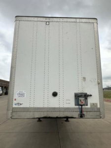Rear view of a white Wabash trailer with doors closed, showing maintenance labels and a connection box, on a paved lot.