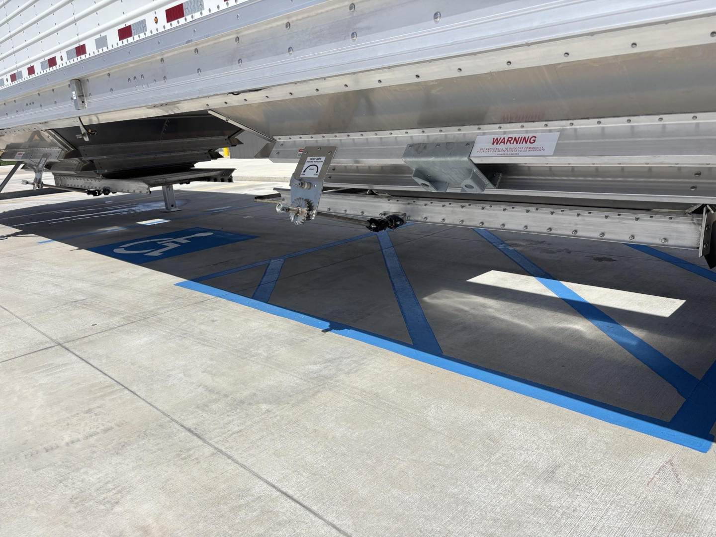 Trailer side view of a 2026 Stoughton 43' Alum Grain Hopper showing metal side panel, ladder, and warning label, with blue parking lines beneath.