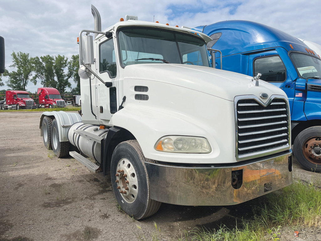 Mack CX613 daycab truck in a lot showing front grille, bumper, and dual rear axles with white cab and chrome accents.