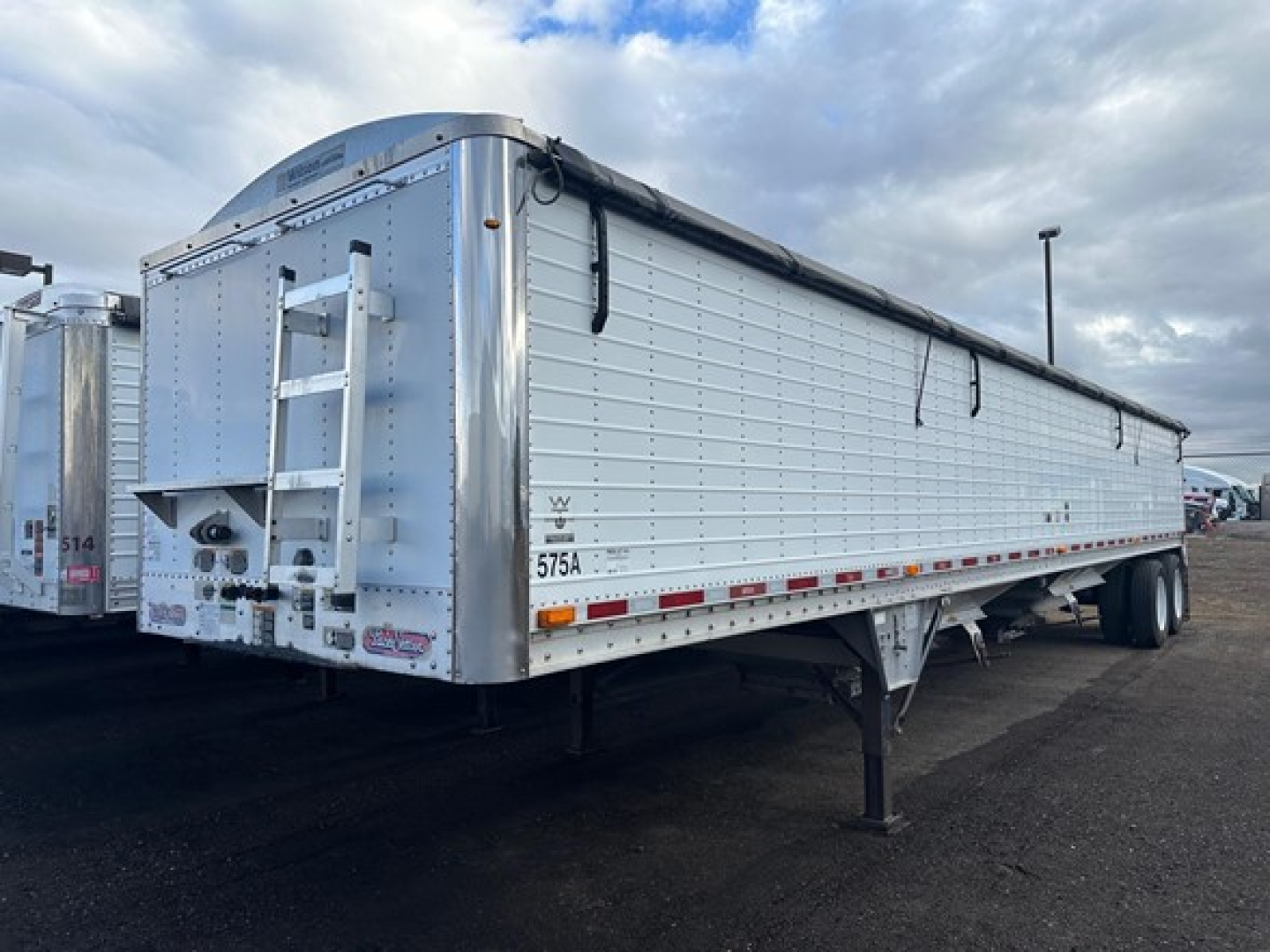 Trailer side view showing a white Wilson Pacesetter hopper trailer with a black tarp and ladder at the rear. Visible text includes '575A' on the side and license plate area at back.