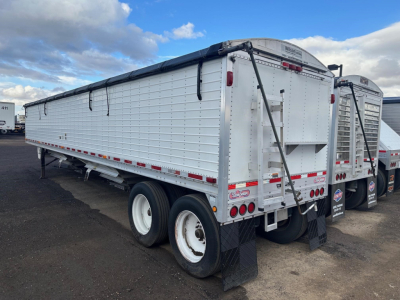 Truck trailer rear view showing dual rear doors, bumper, and dual axle wheels with white rims; the trailer has a metal ribbed side with a black tarp roll and red/white reflective tape along bottom edge.