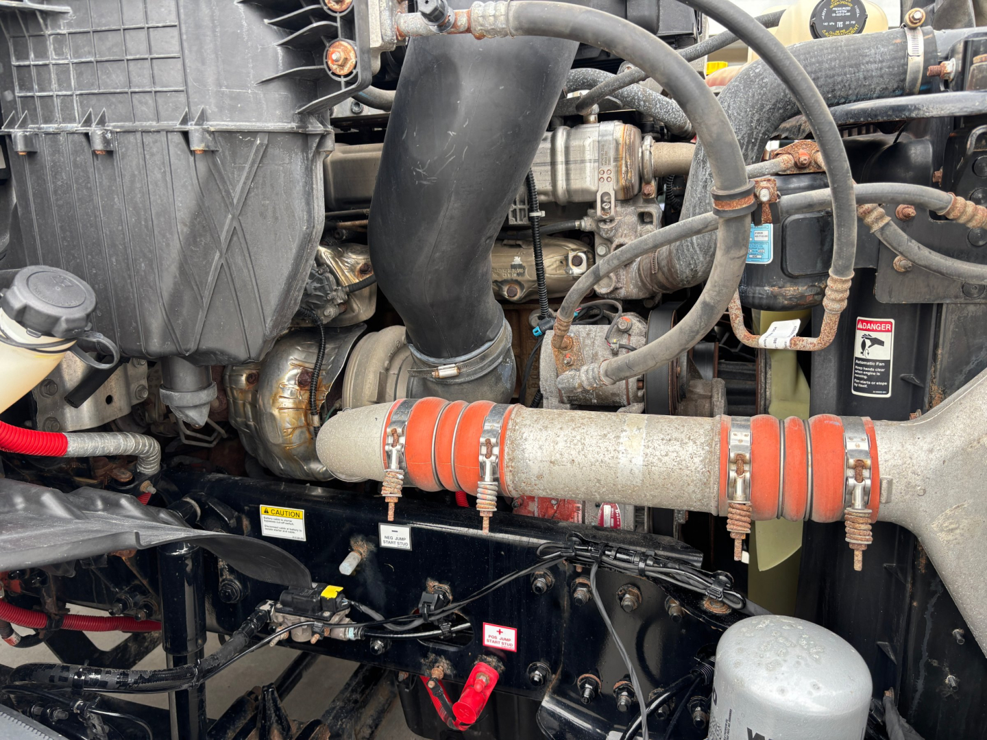 Engine bay view showing hoses, clamps, and a large orange-banded pipe across with metal supports. Visible components include a black air intake housing, tubing, electrical wiring, and a gray cylindrical reservoir.