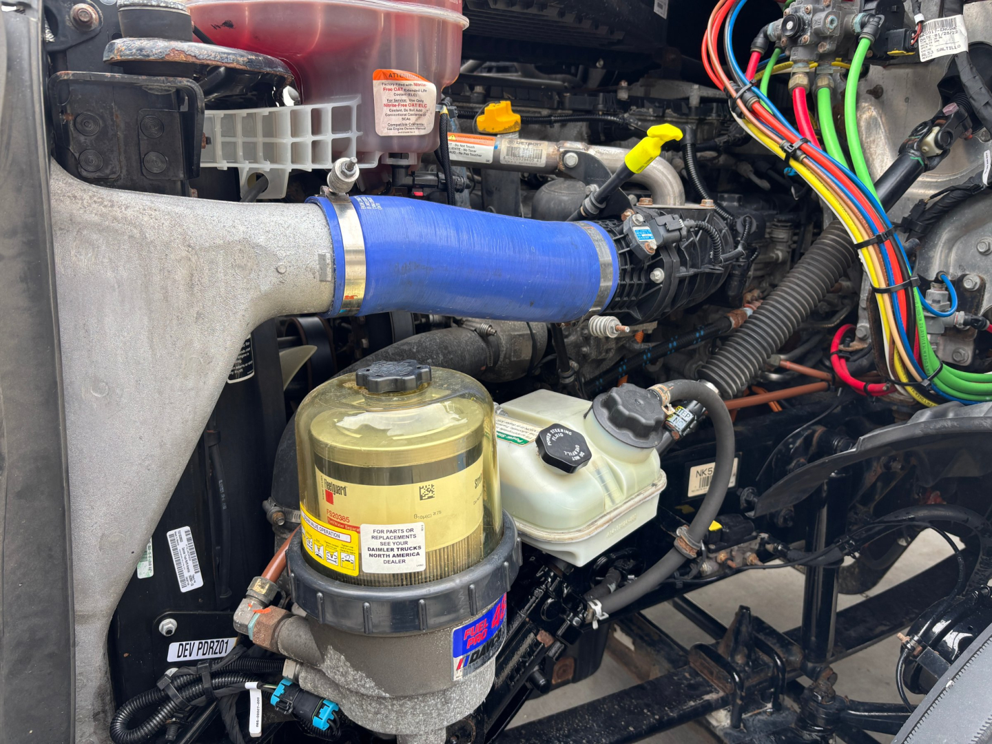 Truck engine bay showing blue silicone intercooler hose, yellow coolant reservoir cap, hydraulic brake fluid reservoir, wiring harness, and various labeled fluid caps.