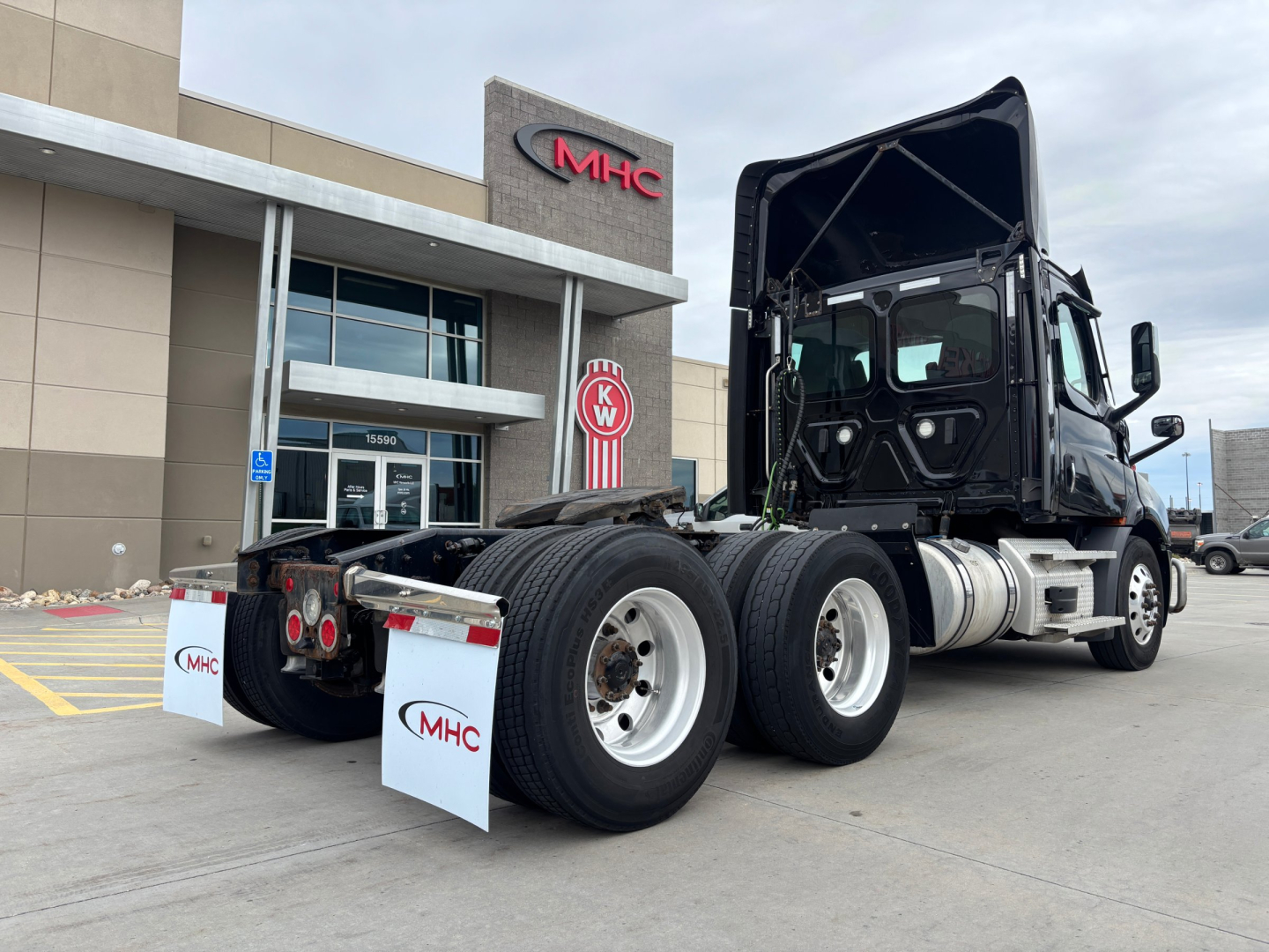Truck chassis of a Freightliner daycab with rear dual-axle configuration, fifth wheel, and dual tires visible; dealership building in background.