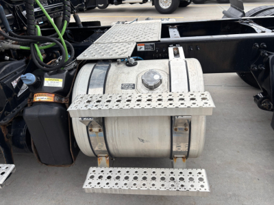 Trucking fuel tank and step panels on Freightliner truck chassis showing cylindrical aluminum fuel tank with perforated steel steps and nearby black battery box; label stickers visible.