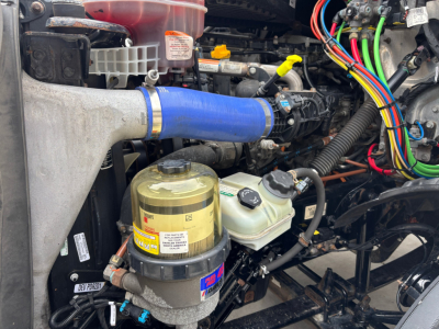 Truck engine bay showing blue silicone intercooler hose, yellow coolant reservoir cap, hydraulic brake fluid reservoir, wiring harness, and various labeled fluid caps.