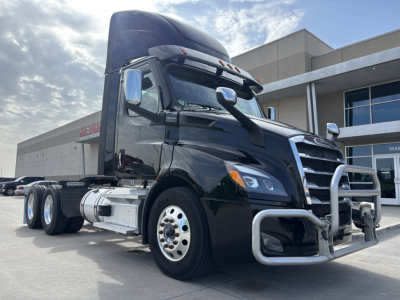 Freightliner daycab tractor showing front grille guard and chrome fuel tanks with dual rear axles and aerodynamic mirrors.