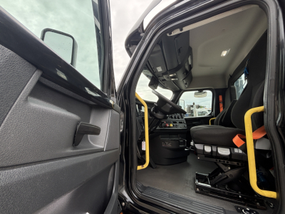 Cab interior of a Freightliner daycab showing steering wheel, driver seat, and entry with yellow grab bars.
