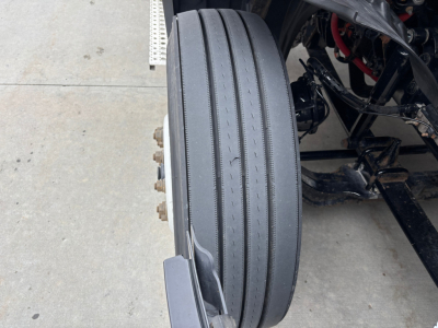 Truck wheel and front suspension showing single solid steer tire with longitudinal tread grooves and surrounding chassis components.