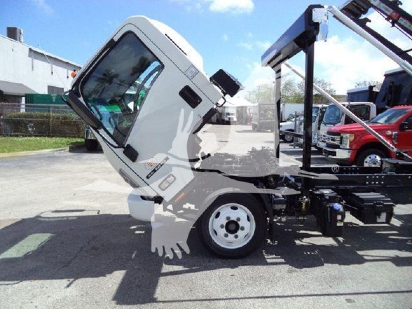 Isuzu NRR cab tilted forward on a roll-off truck setup showing front wheel and cab interior window area with some attached equipment.