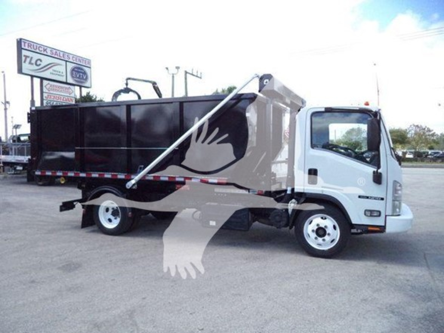 Isuzu NRR roll-off truck with a black dump body, white cab, and crane setup. Visible text includes a sign in the background and reflections; no readable VIN or serials on the vehicle itself.