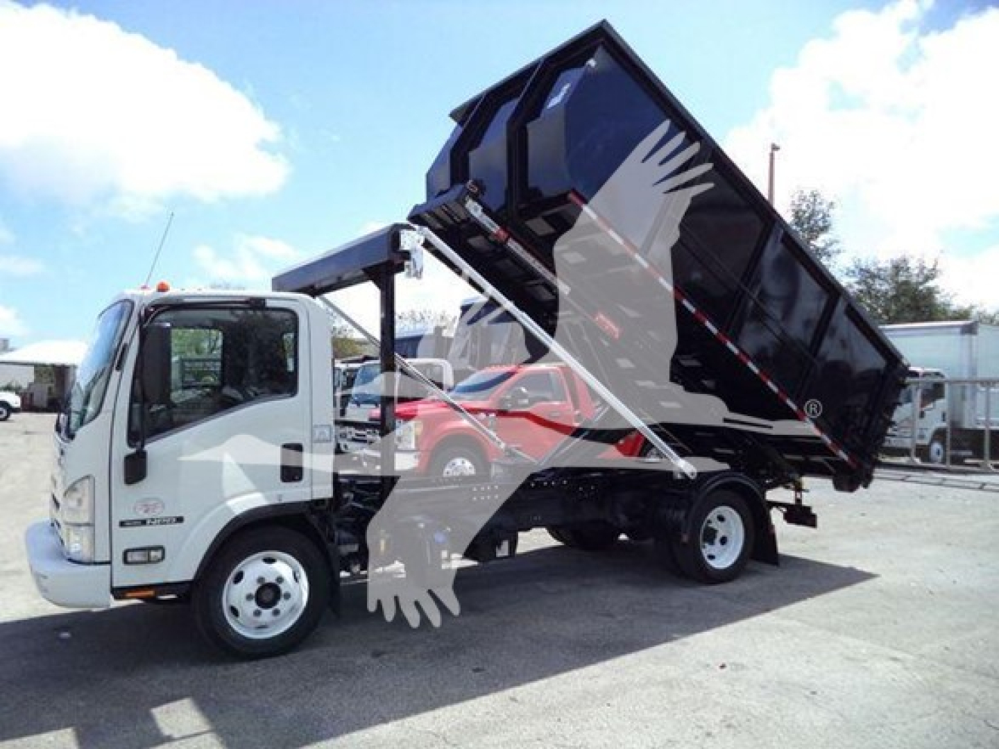 Isuzu NRR roll-off truck with lifted black container in open position; cab front left angle; light grey truck with white wheels; blue sky