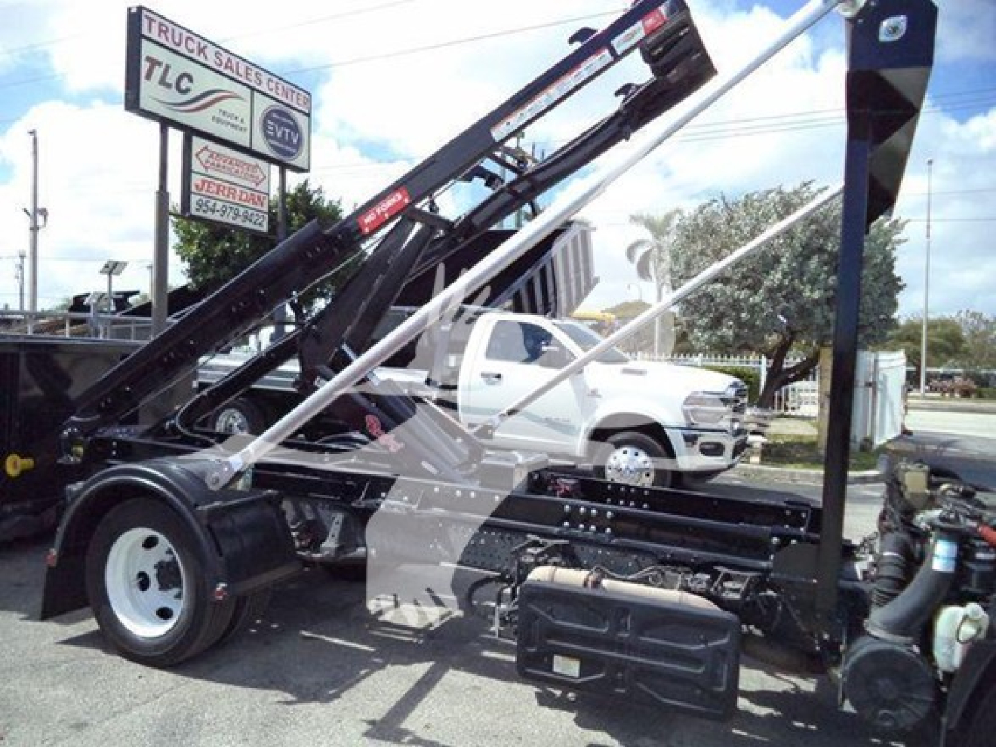 Isuzu NRR roll-off truck chassis with crane arm, showing VIN and stock area in the background; no readable labels on the truck components visible.