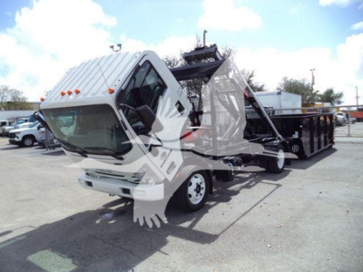 Isuzu NRR truck with roll-off bed tilted, front view showing cab and hydraulic system, stock and lot in background.
